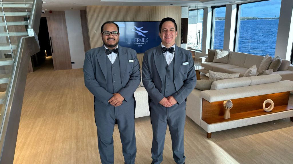 Two smiling butlers aboard in formal grey uniforms stand in the elegant main lounge of the luxury  Galapagos Mega Catamaran Hermes, with panoramic ocean views in the background.