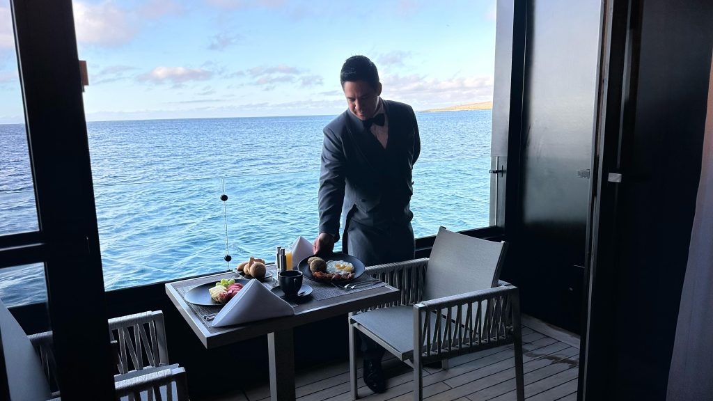One of the 2 butlers in formal uniform serves a gourmet breakfast on a private balcony aboard the luxury Mega Catamaran Hermes, with the serene waters of the Galápagos in the background.