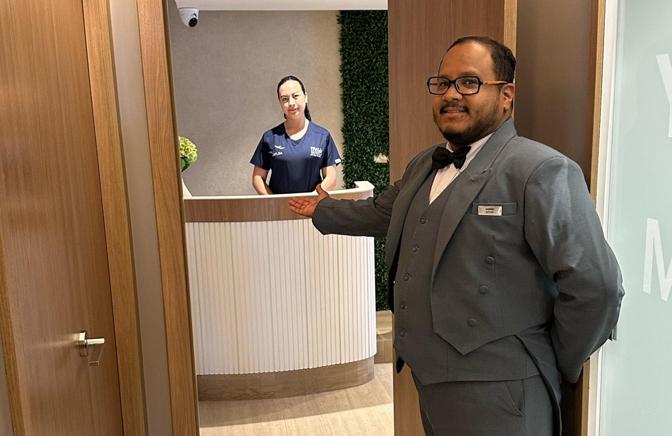 A butler aboard the luxury mega catamaran Hermes escorts a guest to the spa for a personalized massage appointment, where a therapist awaits at the reception.