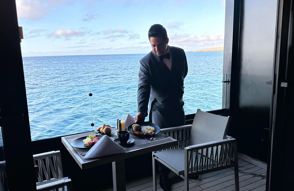 One of the 2 butlers in formal uniform serves a gourmet breakfast on a private balcony aboard the luxury Mega Catamaran Hermes, with the serene waters of the Galápagos in the background.